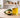 Unlabeled glass bottle of golden avocado oil beside one whole avocado and one halved avocado on a light neutral worktable in bright natural light, with a softly blurred modern workshop background.