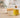 Unlabeled glass bottle of golden chamomile oil beside a handmade soap bar and a few chamomile flowers on a light neutral worktable in bright natural light, with a softly blurred modern workshop background.