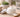 Open glass jar of white facial cream on a textured wooden surface beside a wooden spatula with a dollop of cream, a folded linen towel, a small potted succulent, and a blurred glass of water in warm morning light.