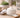 Open glass jar of white facial cream on a textured wooden surface beside a wooden spatula with a dollop of cream, a folded linen towel, a small potted succulent, and a blurred glass of water in warm morning light.