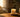 Warm sunlight pours into a cozy living space, illuminating a lit candle on a rustic wooden table beside soft textiles and greenery, creating a calm and inviting atmosphere.