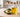 Unlabeled glass bottle of golden avocado oil beside one whole avocado and one halved avocado on a light neutral worktable in bright natural light, with a softly blurred modern workshop background.