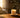 Warm sunlight pours into a cozy living space, illuminating a lit candle on a rustic wooden table beside soft textiles and greenery, creating a calm and inviting atmosphere.