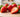 Close-up of several fresh strawberries on a light wooden surface in bright natural light, with the image emphasizing the tiny seeds and texture on the fruit’s surface against a softly blurred background.
