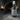 A matte black spray bottle labeled 'Hallowed Ground' set against a dark, atmospheric background of a stone archway covered in cobwebs with candles lit in the distance.
