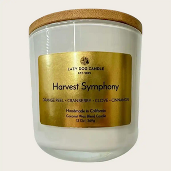 A 13 oz coconut wax candle in a white glass jar with a light brown bamboo lid, featuring a metallic gold label that reads 'Harvest Symphony' by Lazy Dog Candle.