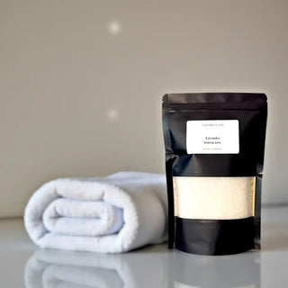Black packaging of lavender bath salts with a white towel on a reflective surface