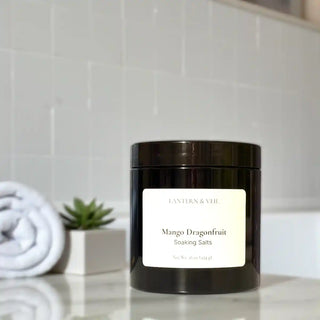 Black jar labeled 'Mango Dragonfruit Soaking Salts' on a bathroom counter with towels and a plant in the background.