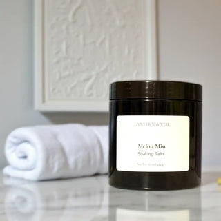 Black jar of 'Melon Mist' soaking salts on a marble surface with a white towel in the background.