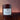 A dark wine glass jar candle with a white label reading 'Midnight Harvest', set on a dark wooden surface against a warm cranberry background.