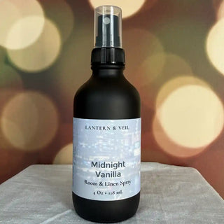 Black bottle of 'Midnight Vanilla' room & linen spray by Lantern & Veil on a textured surface with blurred lights in the background.