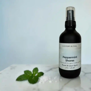 Bottle of 'Peppermint Shores' room and linen spray with a label on a marble surface with peppermint leaves and sea salt.