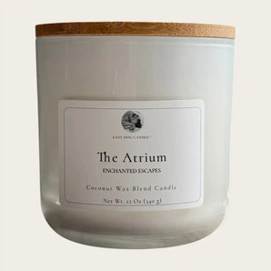 A white jar candle with a light wood lid, featuring a white label that reads 'Lazy Dog Candle The Atrium Enchanted Escapes Coconut Wax Blend Candle'.