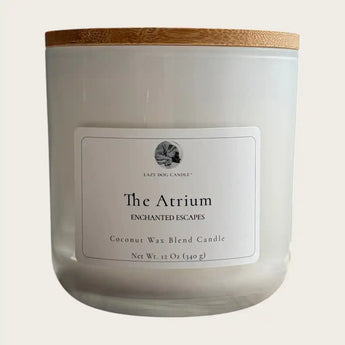 A white jar candle with a light wood lid, featuring a white label that reads 'Lazy Dog Candle The Atrium Enchanted Escapes Coconut Wax Blend Candle'.