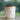 A white jar candle with a light wood lid sits on a light wood surface against a blurred green background with raindrops. The front label reads 'The Atrium' 