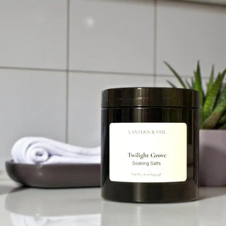 Jar of 'Twilight Grove Soaking Salts' by Lantern & Veil on a bathroom counter with a towel and plant in the background.